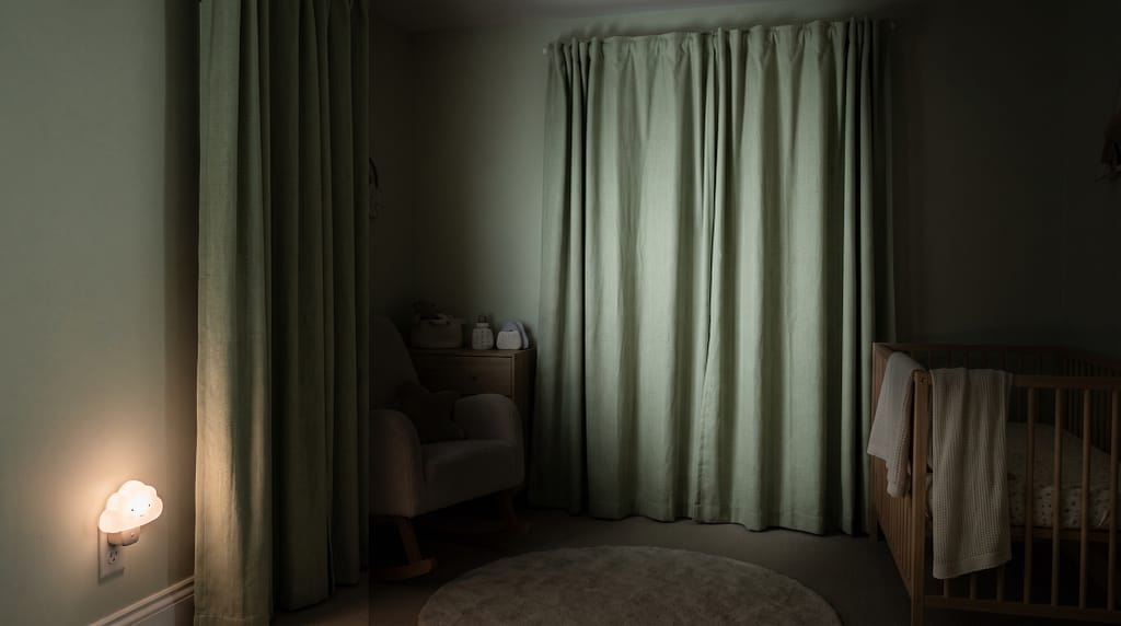 Soft nursery with blackout curtains in pale sage green, cot visible in the background in near-complete darkness